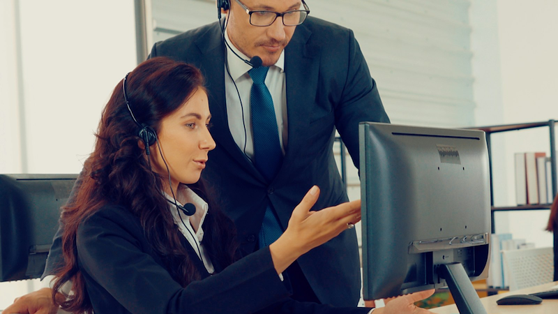 An employee wearing a headset gestures to her computer, with her supervisor looking over her shoulder.