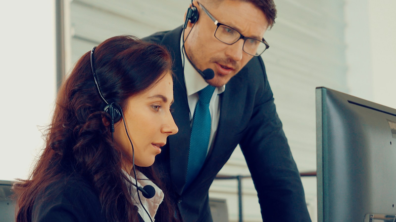 A boss leans over an employee's computer desk while speaking to her as they both wear headsets.