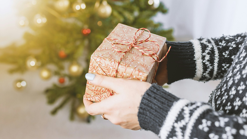 A close-up of a woman's hands holding a festively wrapped gift in front of a decorated Christmas tree.