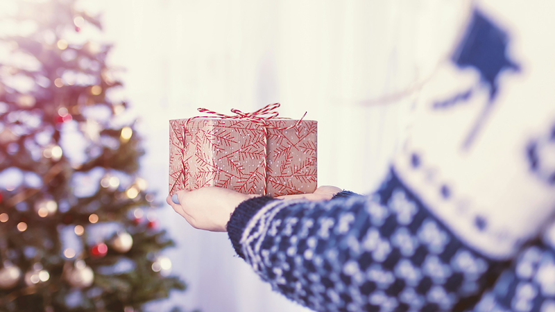 Close-up of a woman approaching a Christmas tree with a festively wrapped gift balanced on her hands.