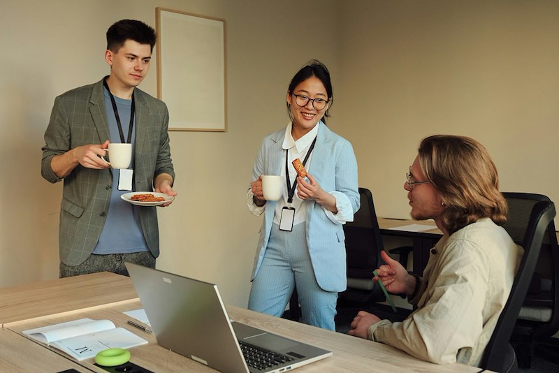 Three employees stand around a desk drinking coffee