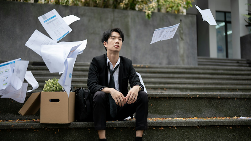 A man in a suit with a loosened tie sits on steps outside while papers flutter around him, a box of belongings next to him.