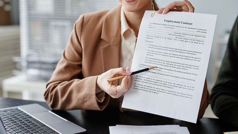 Woman holds an employment contract and points to it with a pen while seated beside a laptop.