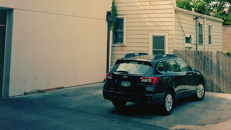 A black car parked in front of a fence and beside a cream-colored building.