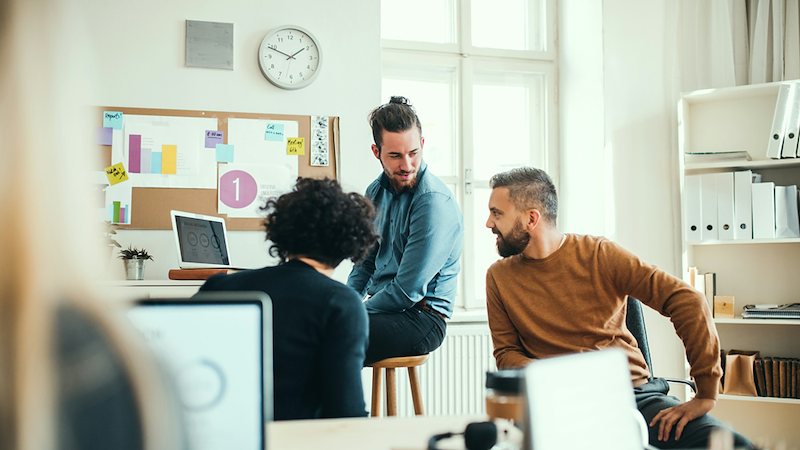 A group of three colleagues sit and talk with one another in an open-plan office.