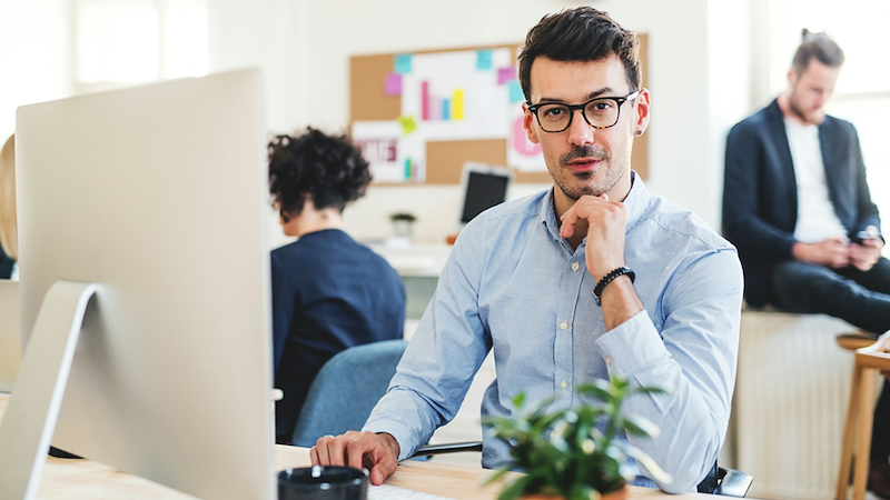An employee rests his head on his chin while sitting at his desk, with coworkers in the background.