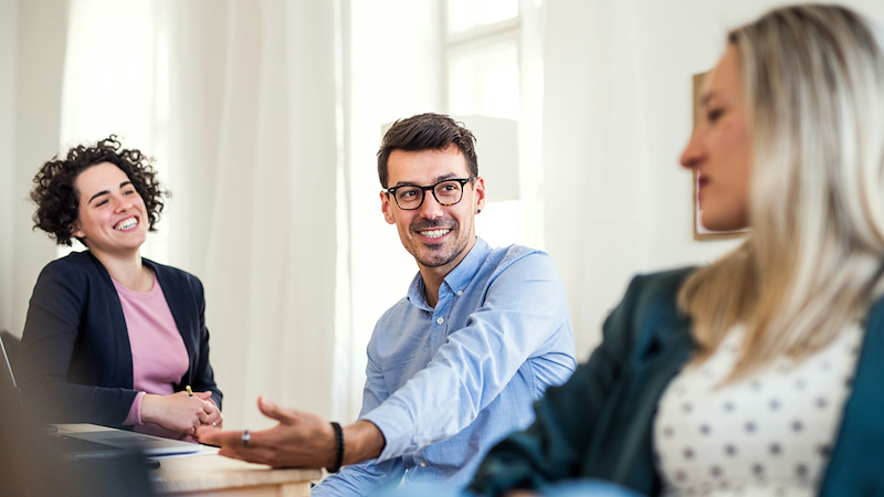 Three coworkers smile as they sit together and discuss something.