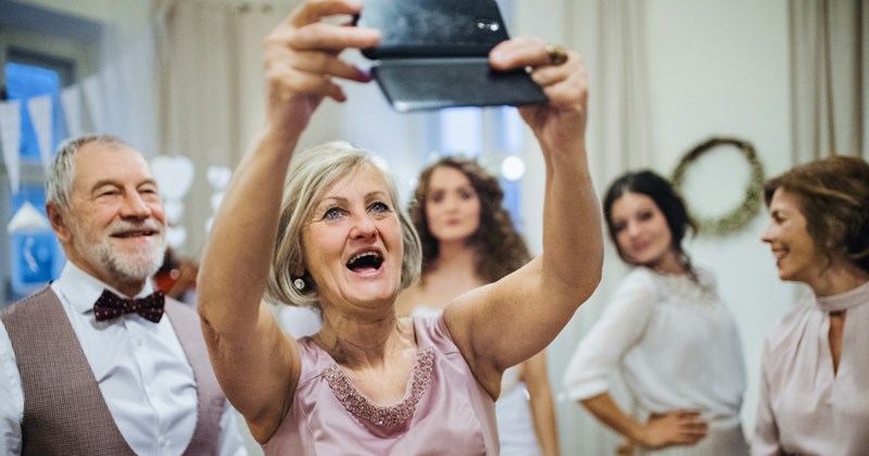 An older female wedding guest smiles as she takes a selfie from above, with the bride and other guests in the background
