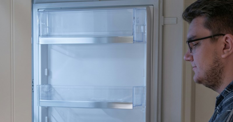 A man wearing glasses looks down into an open refrigerator 