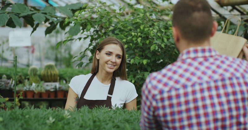 A woman working in a greenhouse smiles at a man holding a paper bag across a table full of plants