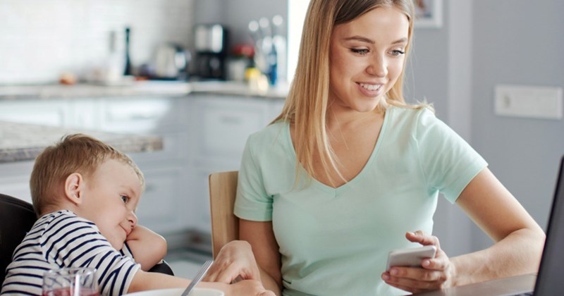 A mom looks at her laptop and holds her cellphone while a young child sits beside her