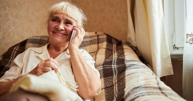 An older woman sitting in an armchair smiles as she calls somebody on her cellphone