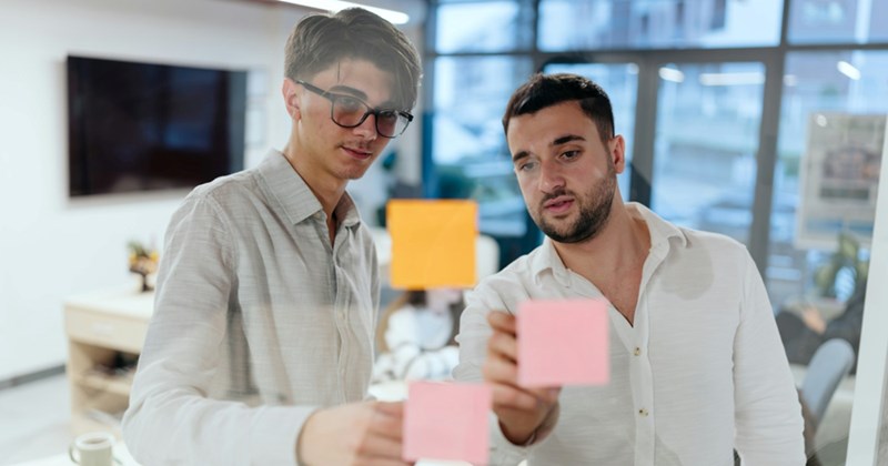Two men stand next to each other in a meeting room, examining two Post-it notes stuck to the window