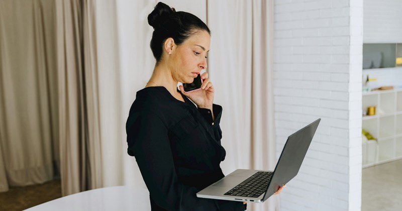 A standing woman dressed in black takes a call on her cell phone while holding her laptop
