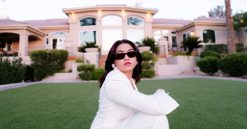 Rich mom wearing a white suit and sunglasses sits on the lawn in front of her mansion 