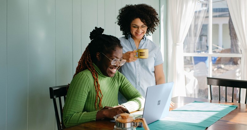 A woman holding a mug stands over a woman working at a laptop on a kitchen table