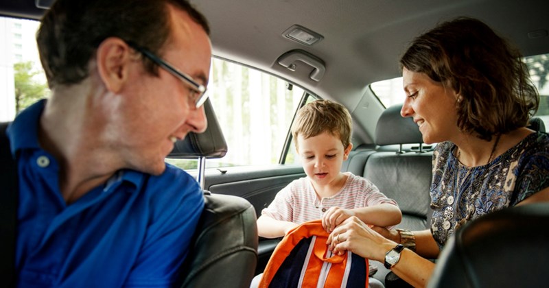A dad in the driver's seat of a car looks towards his son in the back seat, who is being helped by a woman sitting beside him with his backpack