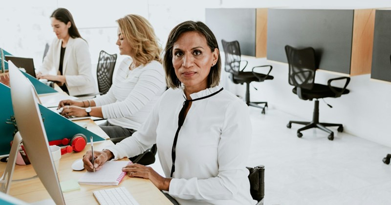 An older woman writes on a notepad in front of her computer, as two colleagues work at their desks behind her