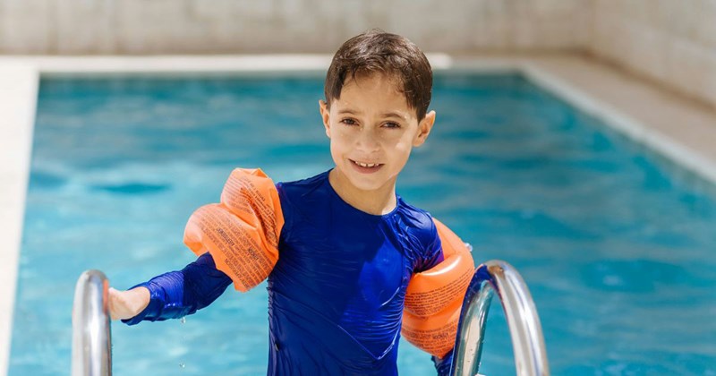 A little boy wearing a swim shirt and floaties emerges from a swimming pool