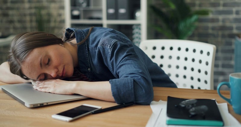 A female employee takes a nap with her head resting on her closed laptop, a notebook, cellphone, and mug beside her