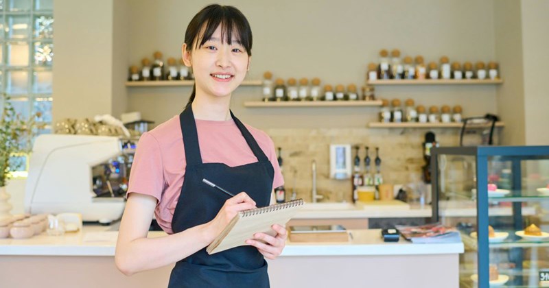 Young Asian bakery employee smiling with a pen and paper, ready to take customer's order