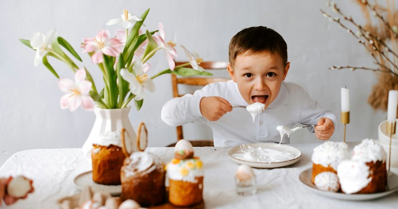 A little boy sitting at a table decorated with plates of a cakes and a bouquet of flowers eats icing from a plate