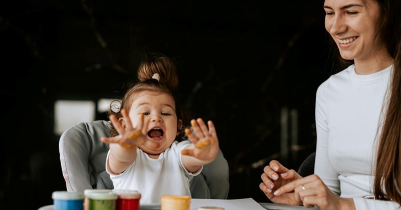 A little girl sitting in a high chair reaches out to the finger paints in front of her while her mom sits beside her, smiling