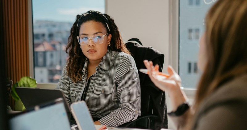 A female employee sitting at a meeting table with a tablet looks towards a female coworker who is talking