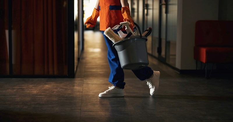 The legs of a woman walking along the floor of an office at night while holding a bucket of cleaning products