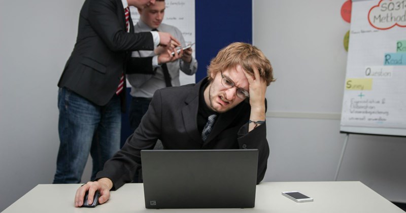 A frustrated employee works at his desk, while his micromanaging manager micromanages his coworker