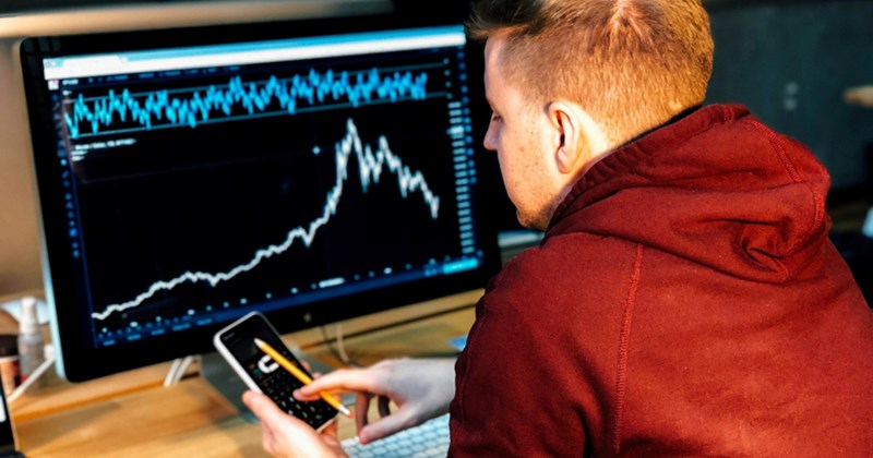 A man sits at a desk with a computer showing a stocks chart, holding his cell phone and pointing at it with a pencil
