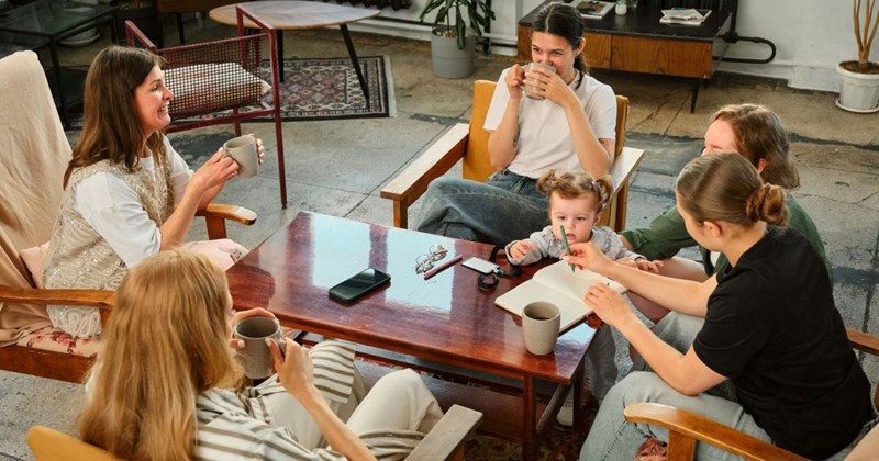 A group of mom friends is drinking tea and talking together while their kids play in another room.