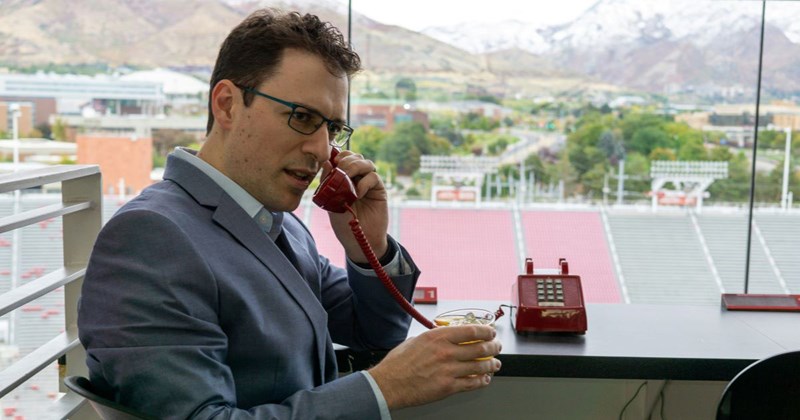 Man in a gray suit talking on the phone while drinking a beverage by a large window