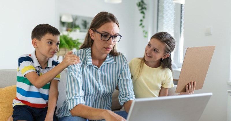 A mom sitting on a couch works from home on her laptop, her son and daughter sat either side of her trying to get her attention
