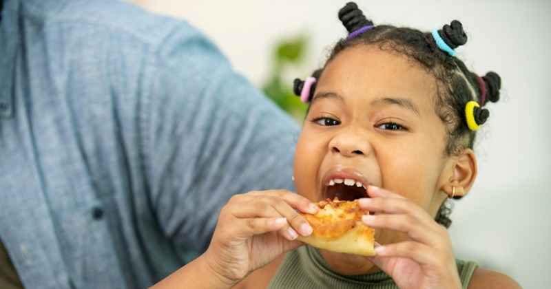 A young female child takes a bite of a pizza slice she is holding with both hands