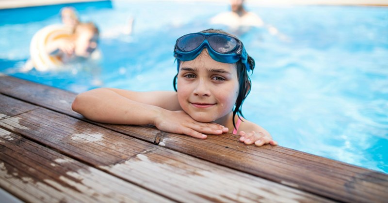 A female child with swimming goggles on her head rests her head and arm on the edge of a swimming pool