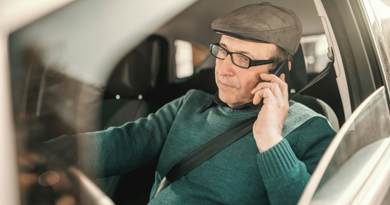 An older man strapped into the driver's seat of a car takes a call on his cell phone