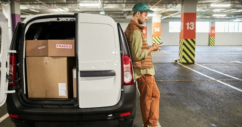 A delivery driver leans against the side of a white delivery van with the back door open, looking at his cell phone