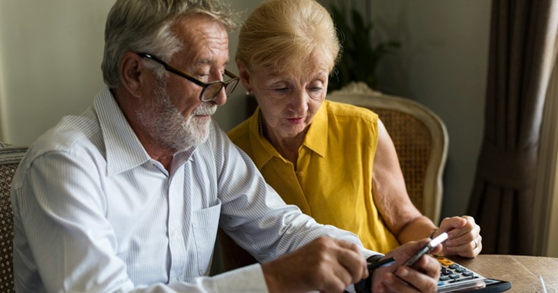 An older couple sits at a table together, looking at something the man is pointing to on his cell phone 