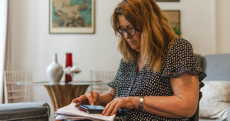 A middle-aged woman wearing glasses sits on a chair in a bedroom, looking at her phone on top of a pile of documents in her lap