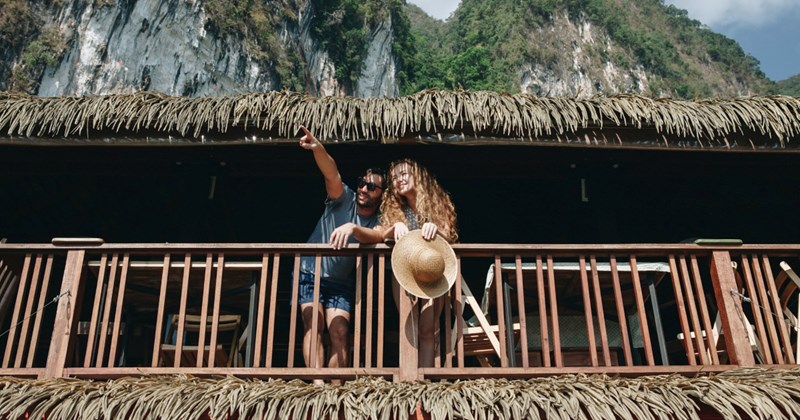 A man and a woman lean over a balcony on a tropical building, with the man pointing to something