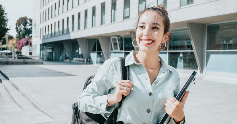 A woman smiles while walking through a college campus, holding a shoulder bag and a laptop