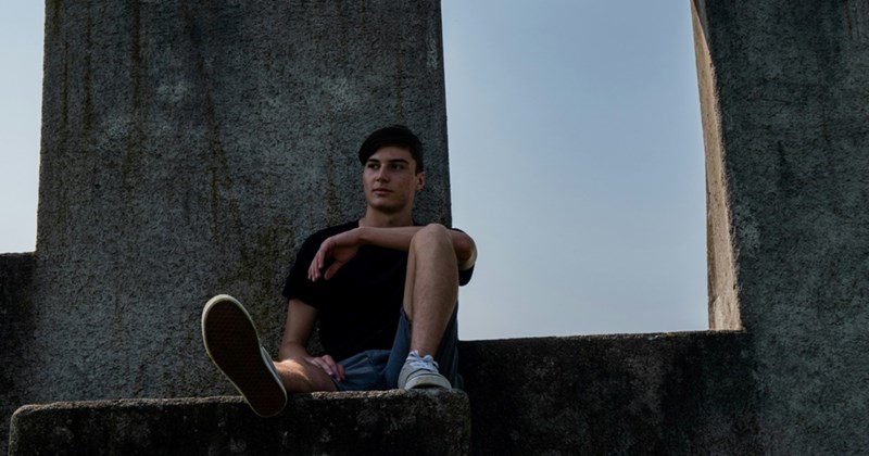 A teenage boy rests his arm on his knee while sitting on a concrete structure outside