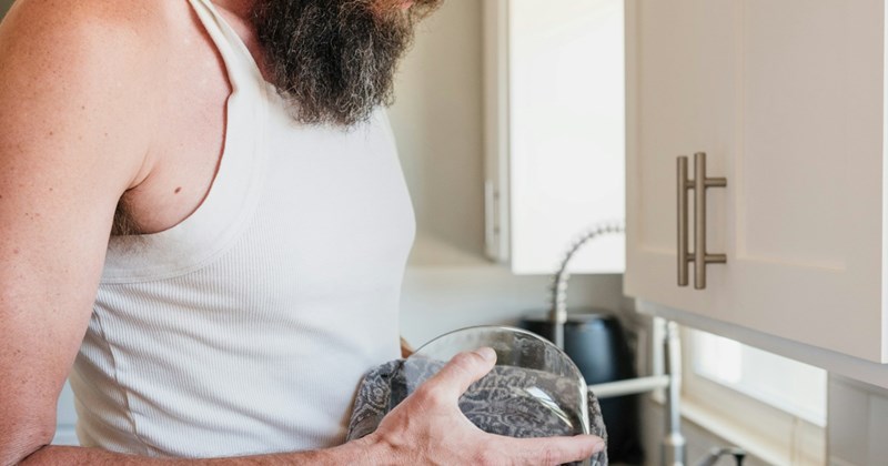 A bearded man wearing a vest dries a glass dish with a dish towel
