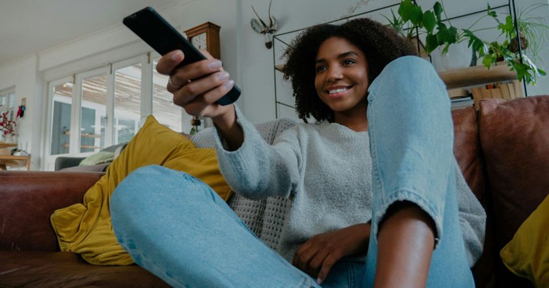 Mixed race woman holding the television remote and changing the channel on the couch while smiling