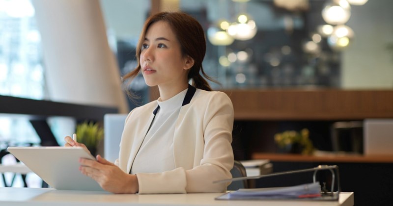 An employee standing at a front desk, thinking about something as she uses a tablet