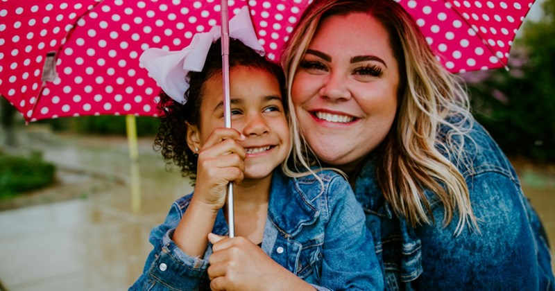 A smiling mom crouches down beside her young daughter, who is holding a pink polka dot umbrella over their heads outside