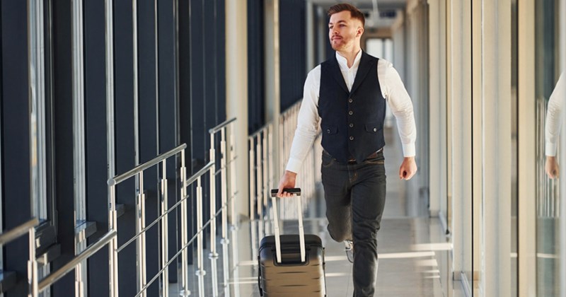 A young male professional runs down an airport hallway, pulling a small suitcase