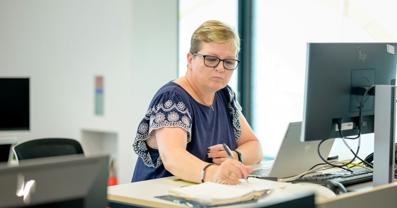 An older female employee sits in front of a computer screen at a desk, taking notes in a notebook