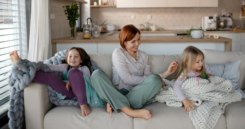 A mom hangs out on a couch with two of her daughters, wrapped in blankets
